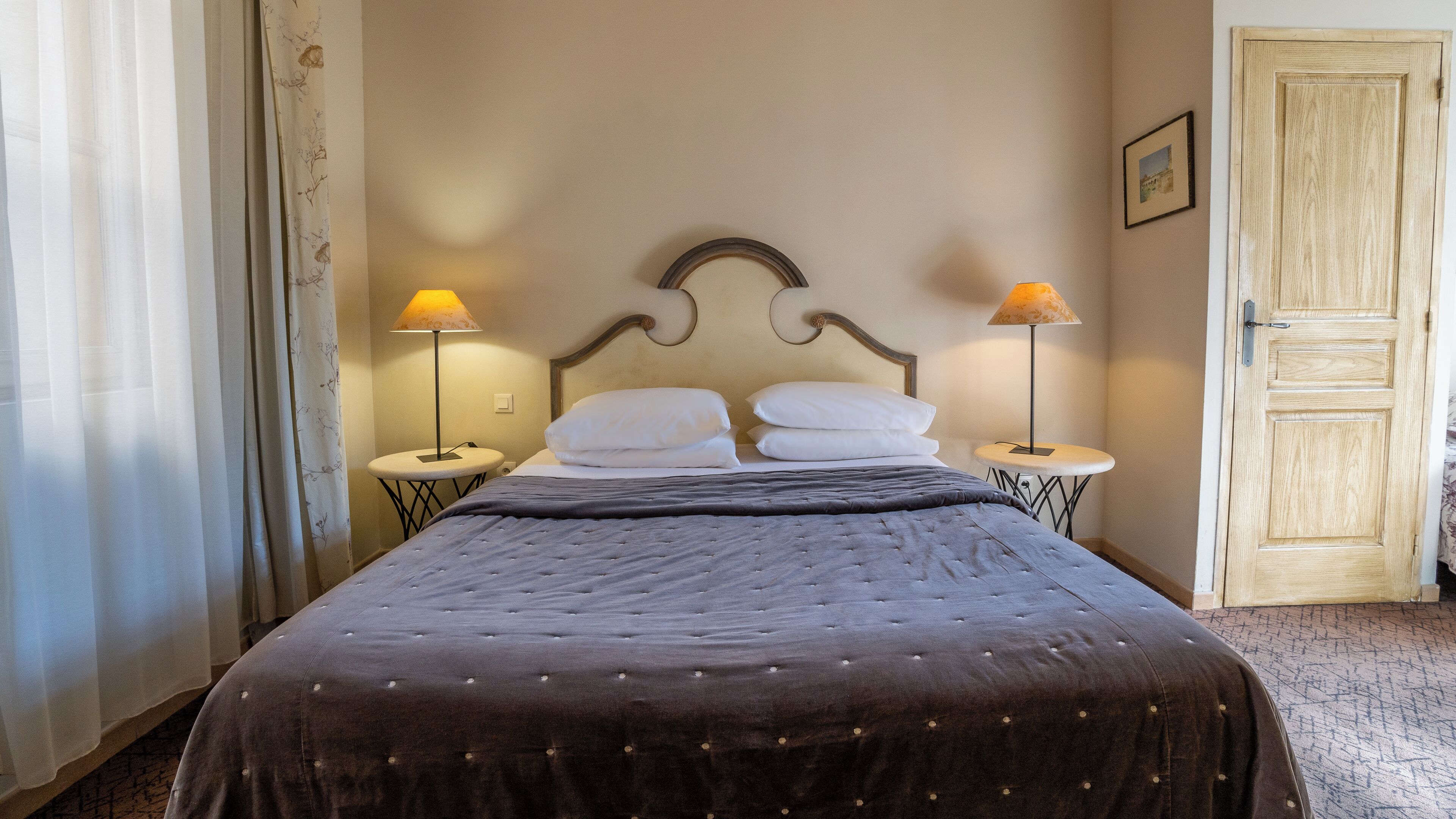 A neatly made bed with a dark duvet and two pillows, flanked by two small bedside tables with lamps, in a room with a wooden door and a framed picture on the wall.