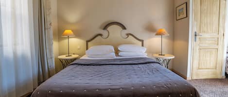 Una cama bien tendida con un edredón oscuro y dos almohadas, flanqueada por dos mesitas de noche con lámparas, en una habitación con una puerta de madera y un cuadro en la pared.