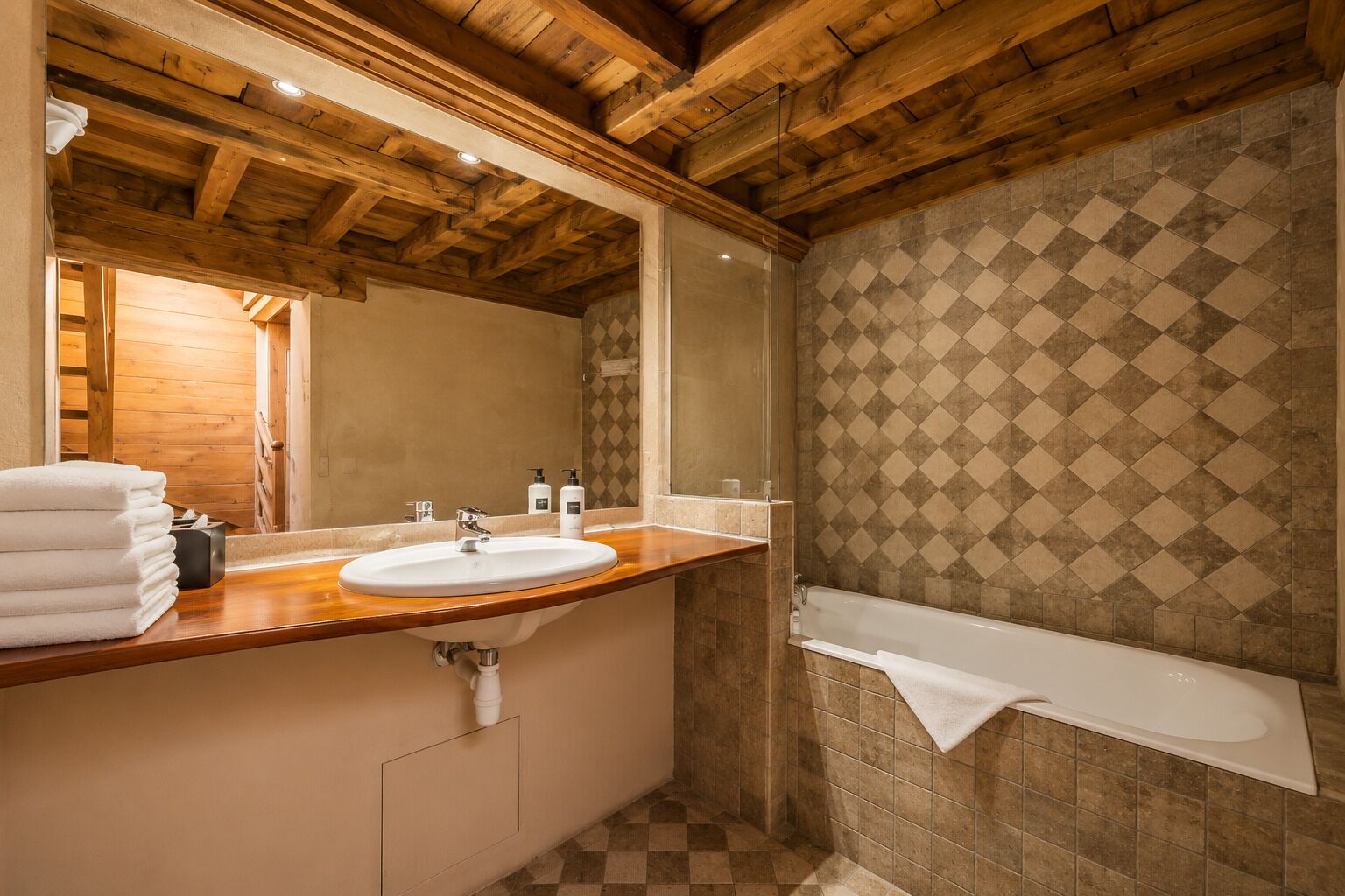 A bathroom with a wooden ceiling, a sink, a mirror, a bathtub, and a patterned wall.