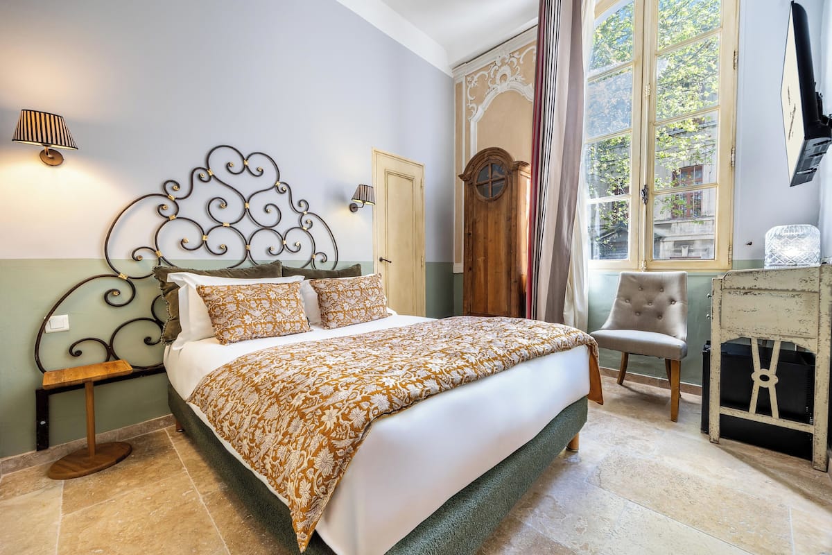 A bed with a patterned bedspread, a bedside table, a chair, a wooden door, and a window with curtains.