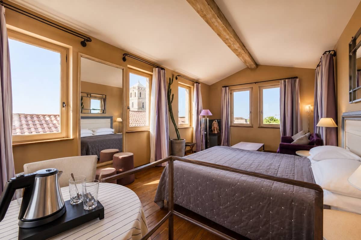 A hotel room with a bed, a desk with a kettle, a chair, and a view of a building outside.