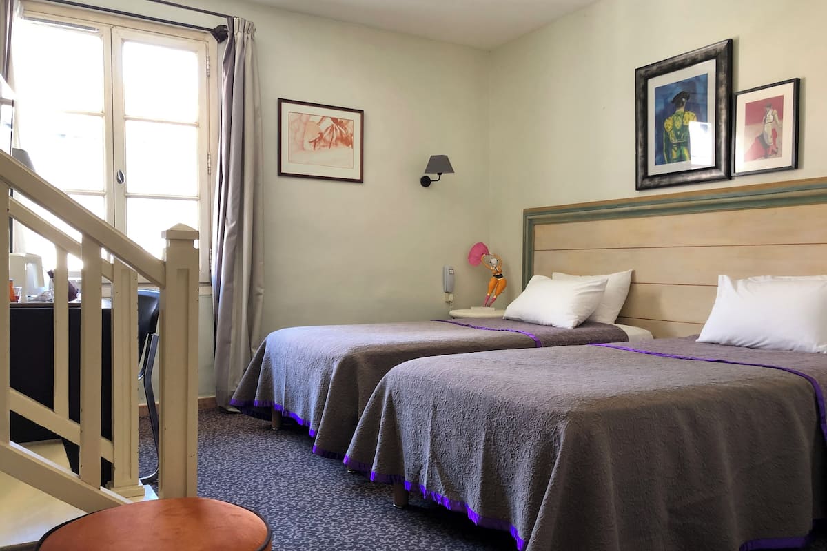A hotel room with two beds, a desk, a chair, a lamp, and a window with curtains.