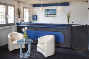 Un espace d’accueil avec un comptoir bleu, deux fauteuils blancs, une table en verre avec un vase et un tableau encadré au mur.