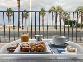 Bandeja de desayuno con pasteles, un vaso de zumo de naranja, una taza y un plato de comida en un balcón con vistas a una playa con palmeras y el océano.