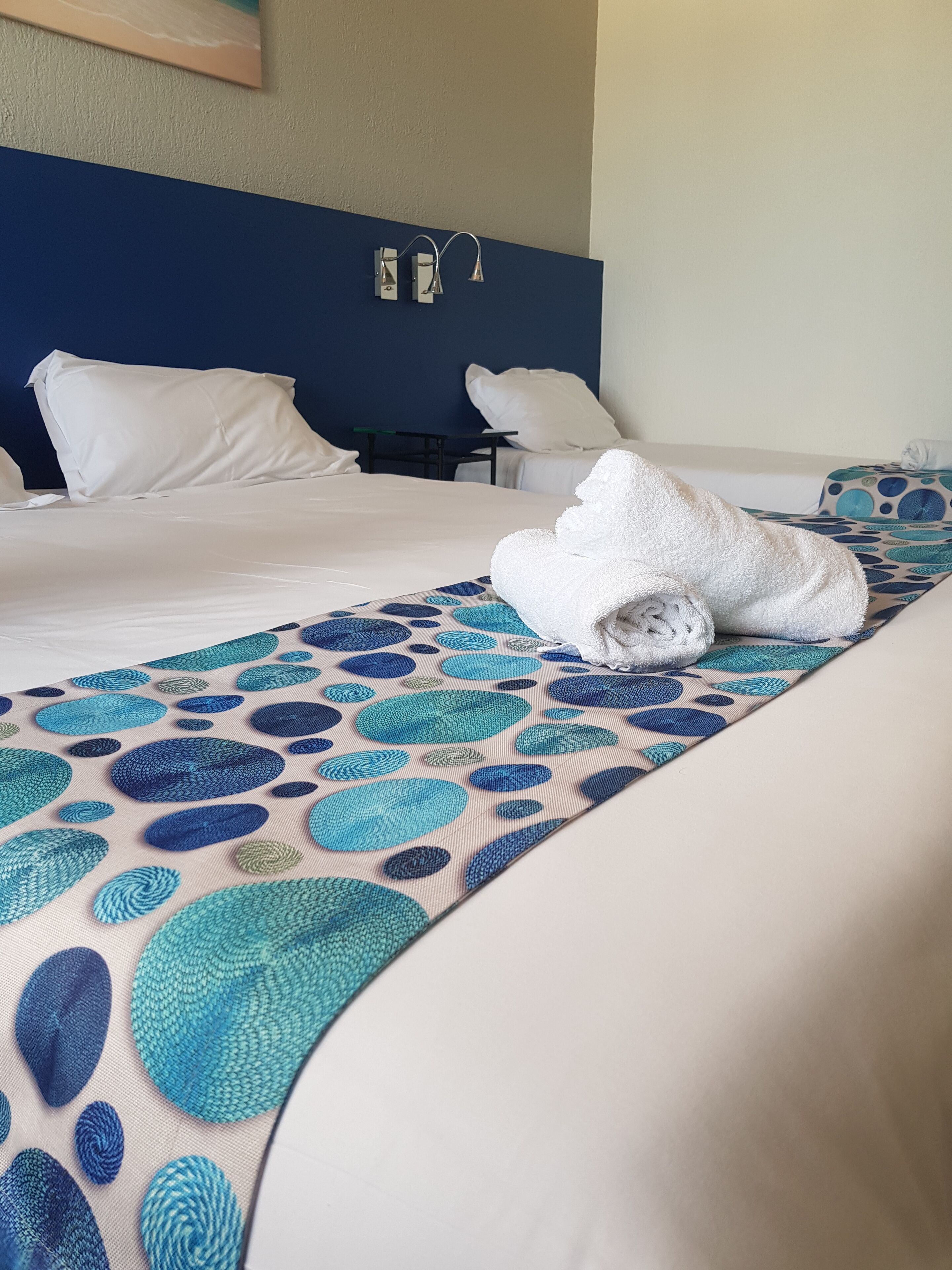 A hotel room with a bed, white pillows, a blue headboard, and a patterned bedspread.