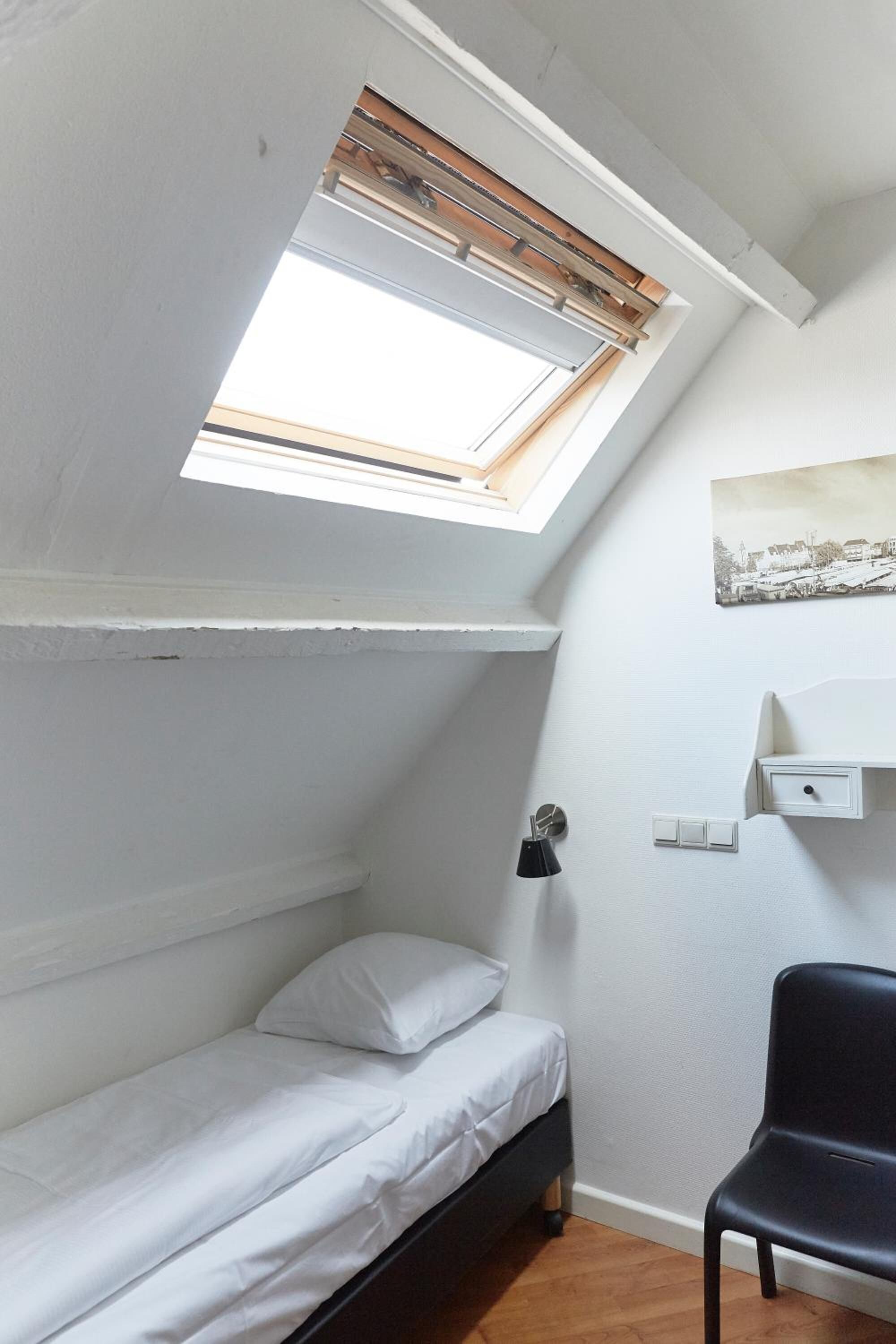 A small, well-lit room with a skylight, a bed, a chair, and a wall-mounted shelf.