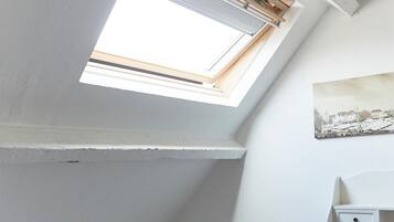 A small, well-lit room with a skylight, a bed, a chair, and a wall-mounted shelf.