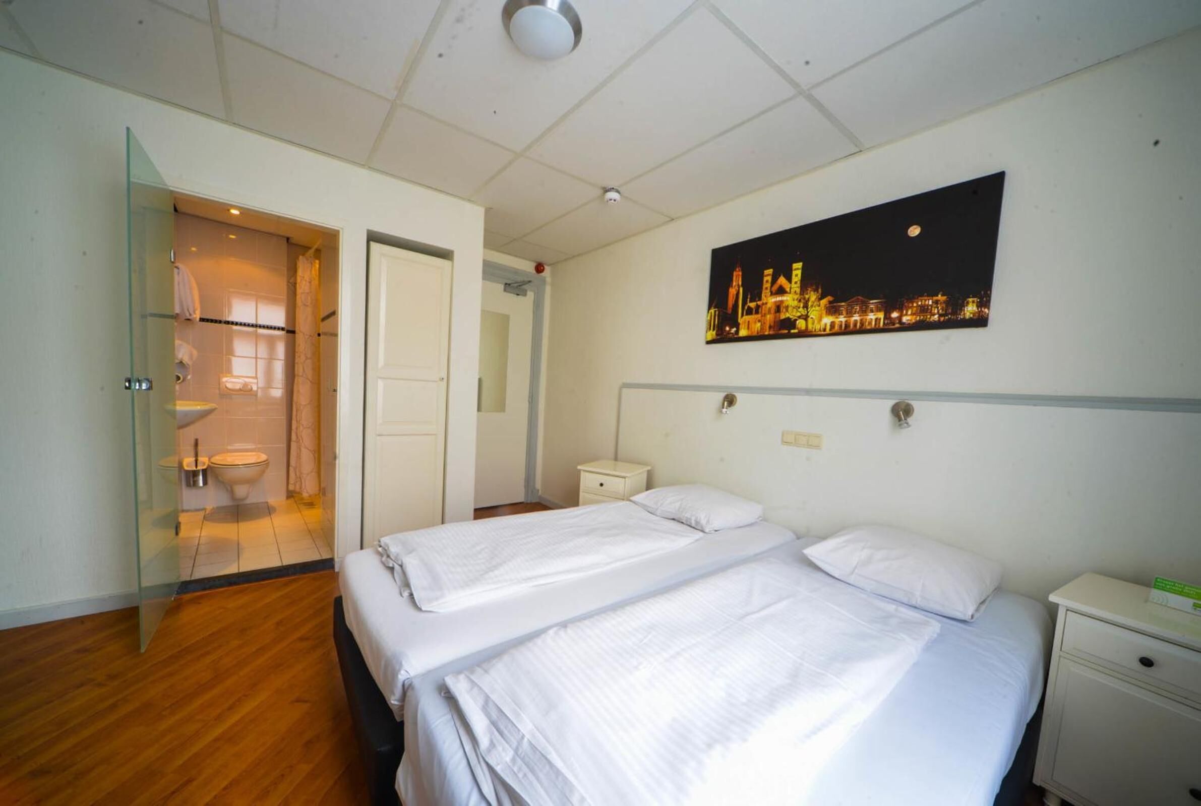 Une chambre d’hôtel avec deux lits, une douche en verre, une table de chevet et un tableau représentant un paysage urbain.