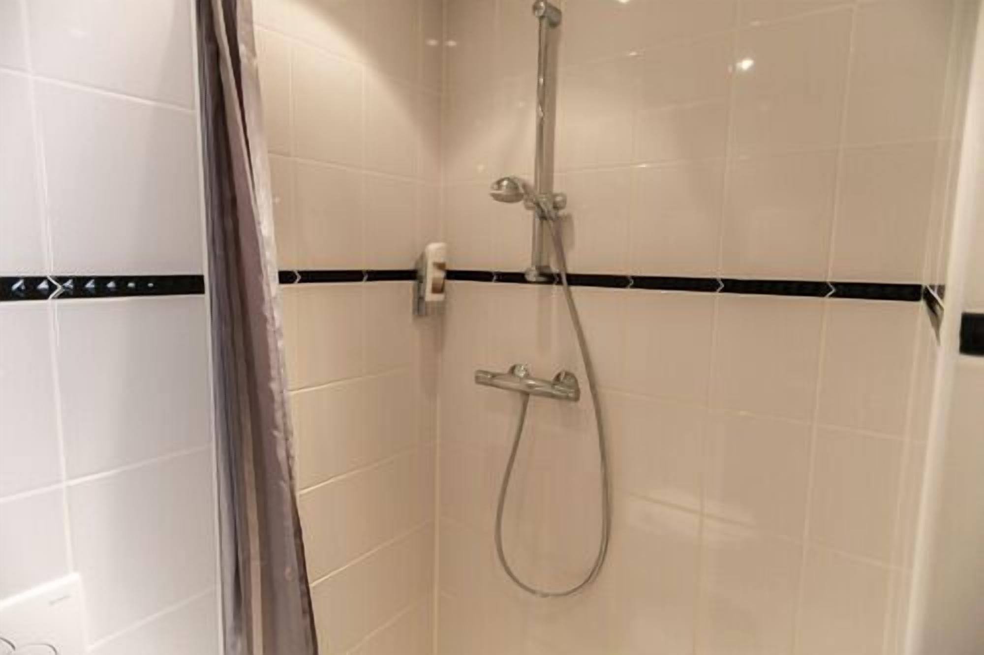Une douche équipée d’un pommeau de douche à main et d’un robinet mural, installée dans une salle de bains carrelée avec une bande horizontale noire sur le mur.