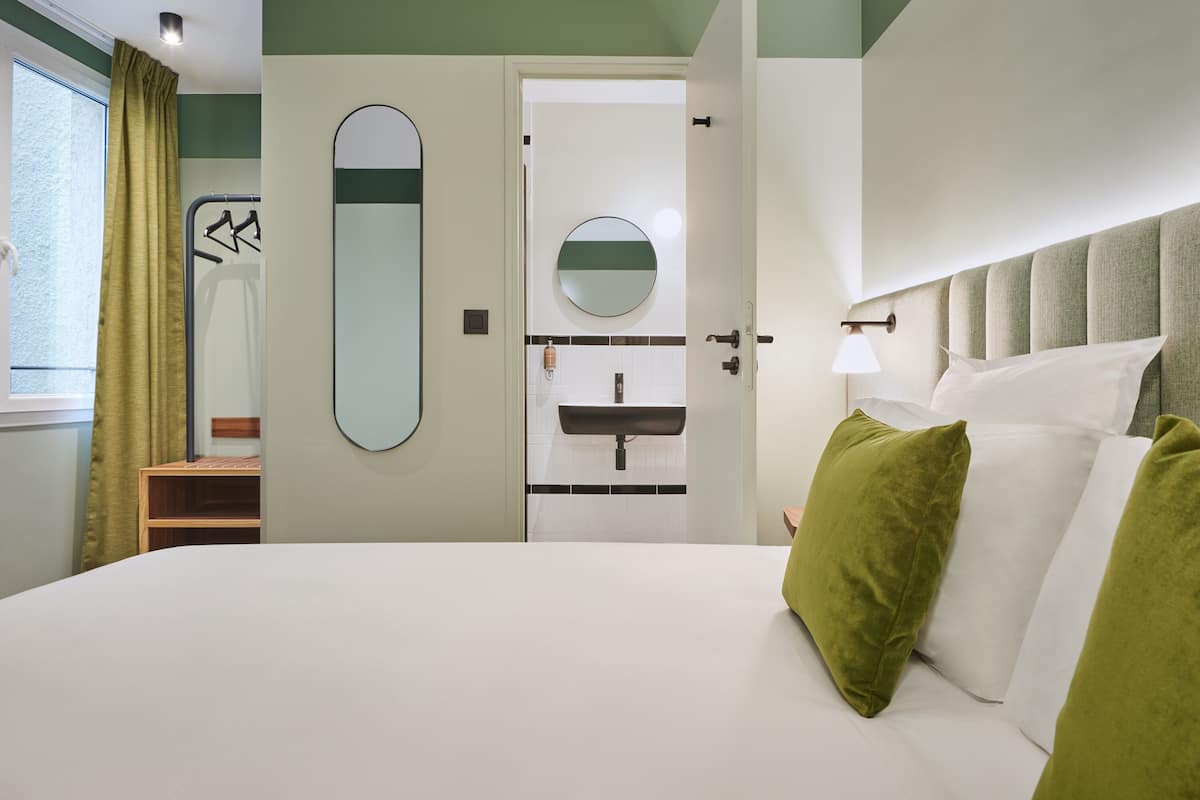 Una habitación de hotel con una cama grande, una mesita de noche, un espejo, un espejo redondo en la pared, una ventana con cortinas y una puerta que da a un baño.