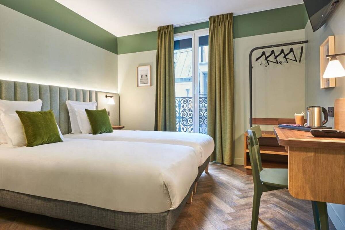 Habitación de hotel con cama, escritorio, silla, ventana con cortinas y lámpara de pared.