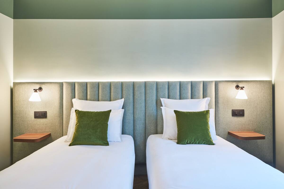 Habitación de hotel con dos camas, cada una con un almohadón verde, y mesitas de noche con lámparas.