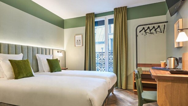 A hotel room with a large bed, a desk, and a chair. There is a window with curtains and a view of a balcony. - Hotel 10 Opera by HappyCulture (Paris)
