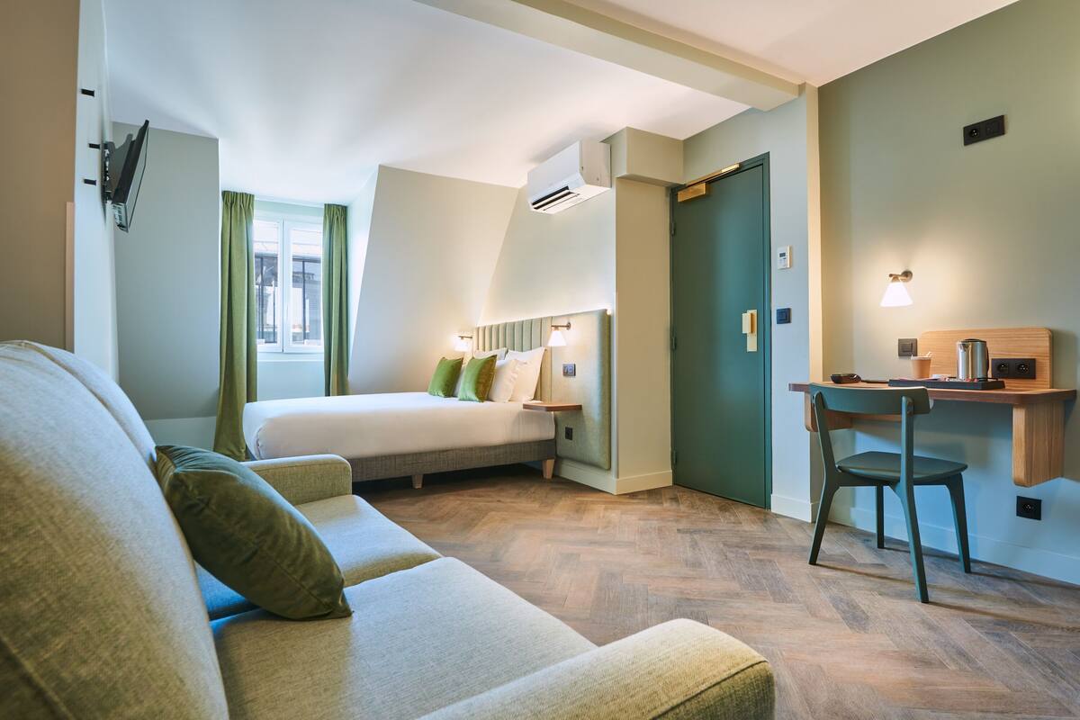 Una habitación de hotel moderna con cama, una zona de estar con sofá, un escritorio con silla y una ventana con cortinas.