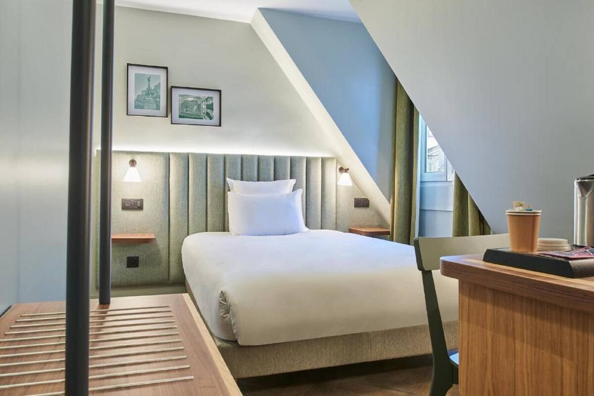 Una habitación de hotel moderna con una cama grande, una mesita de madera y un escritorio con una lámpara. Hay dos luces empotradas en la pared y un cuadro enmarcado sobre la cama.