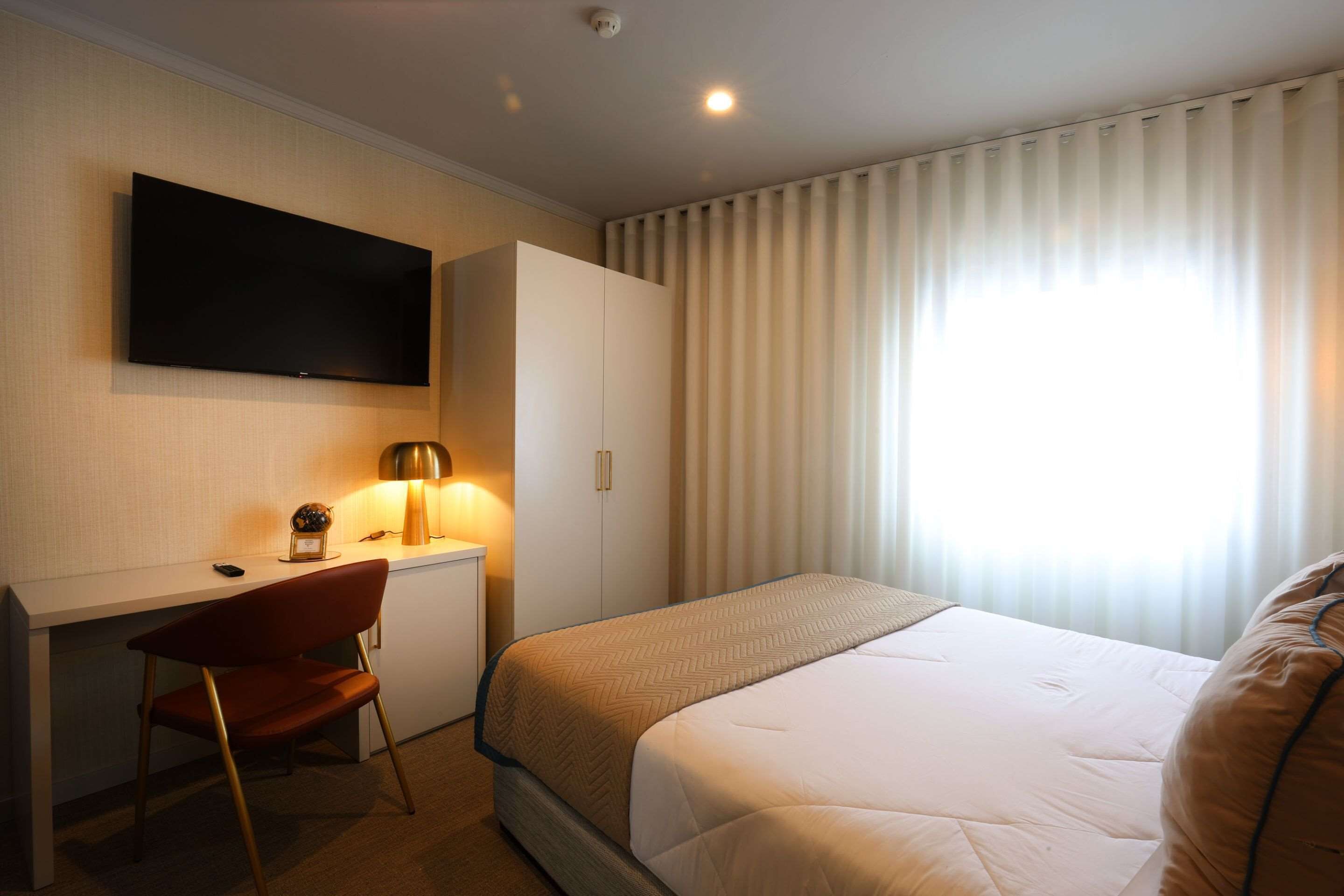 Una habitación de hotel con cama, escritorio con silla, televisión montada en la pared y ventana con cortinas.