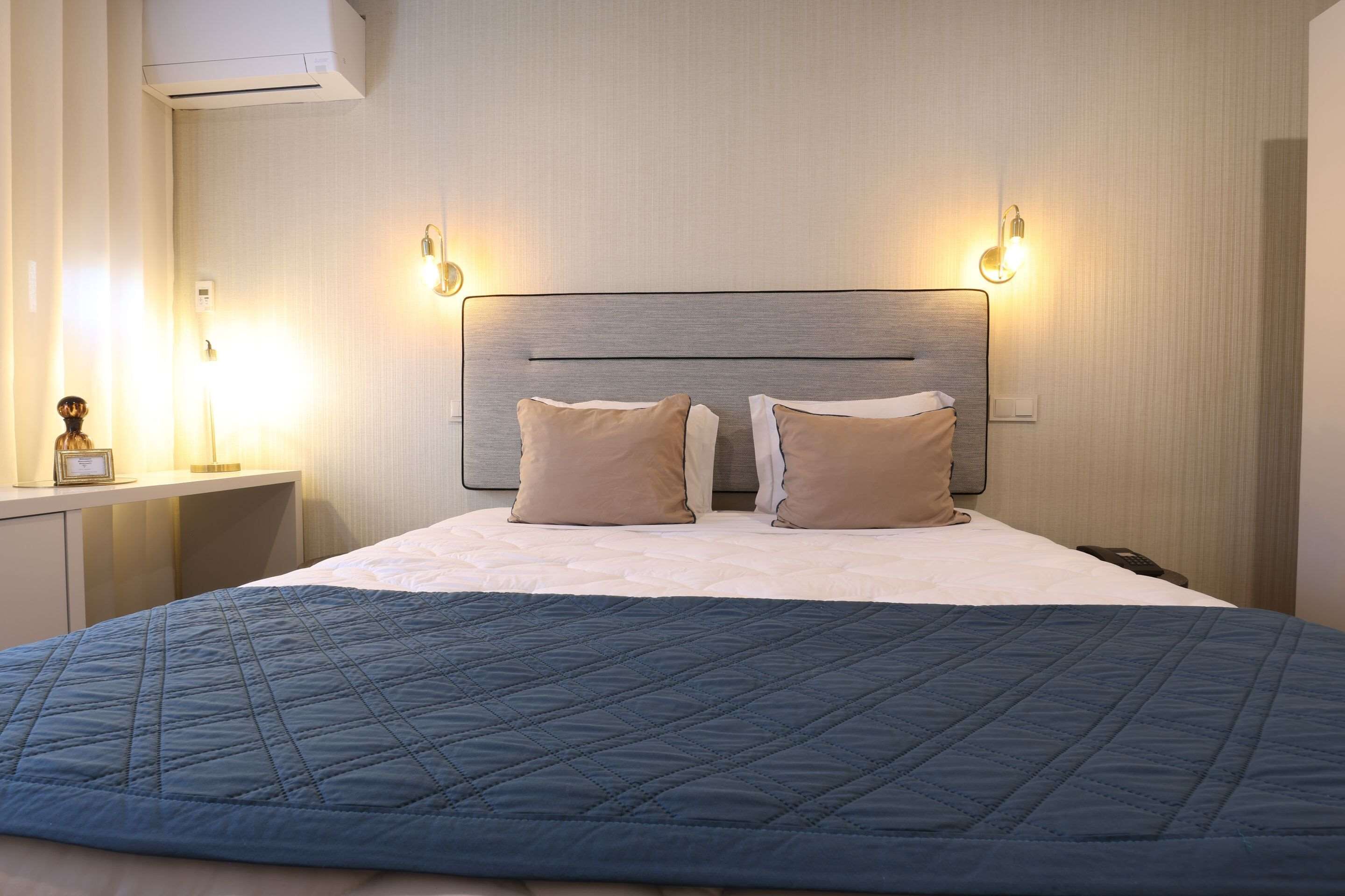 A neatly made bed with a blue quilt, two beige pillows, and a headboard with wall-mounted lights.
