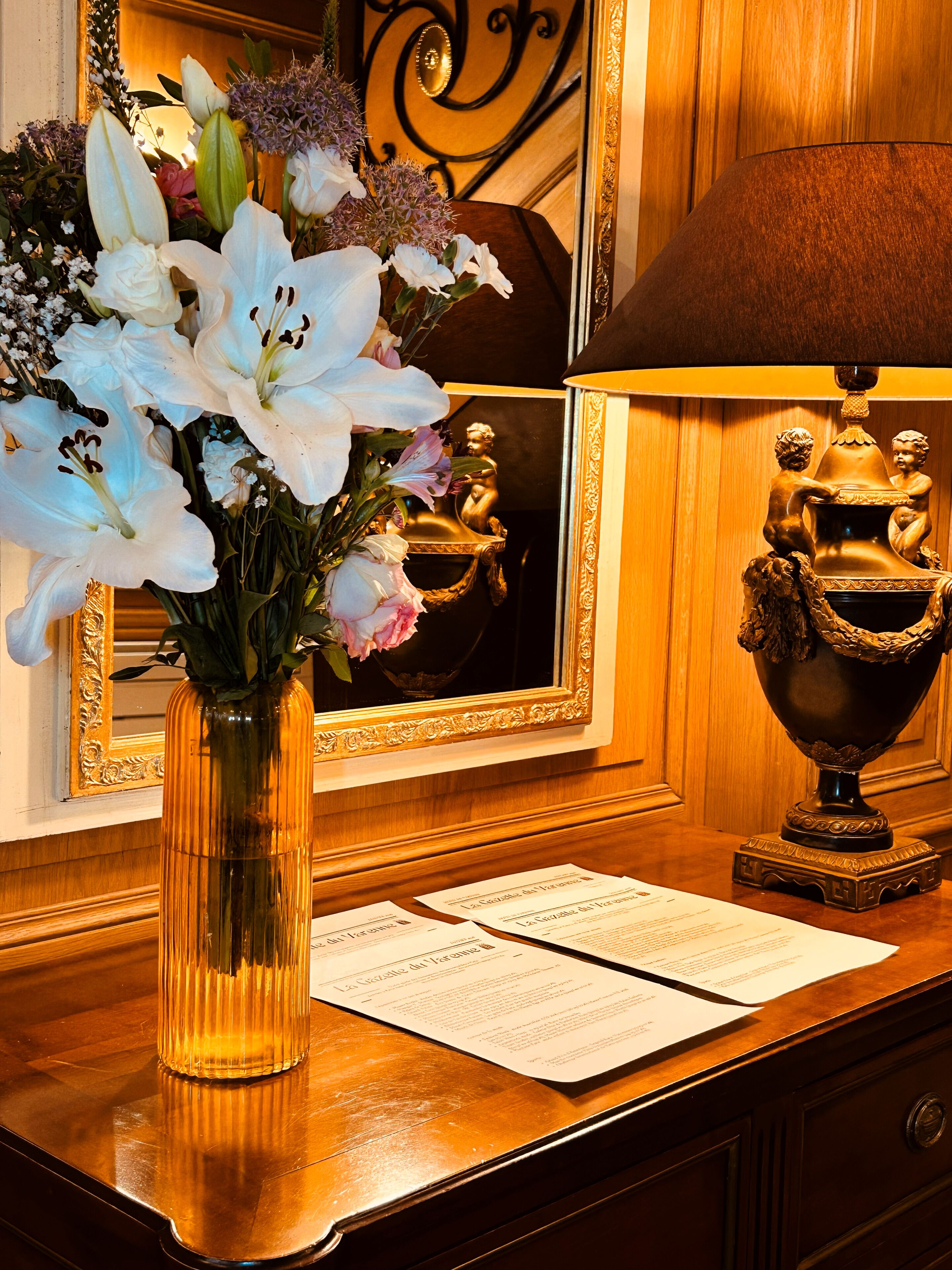 A wooden desk with a vase of flowers, a mirror, and a lamp.