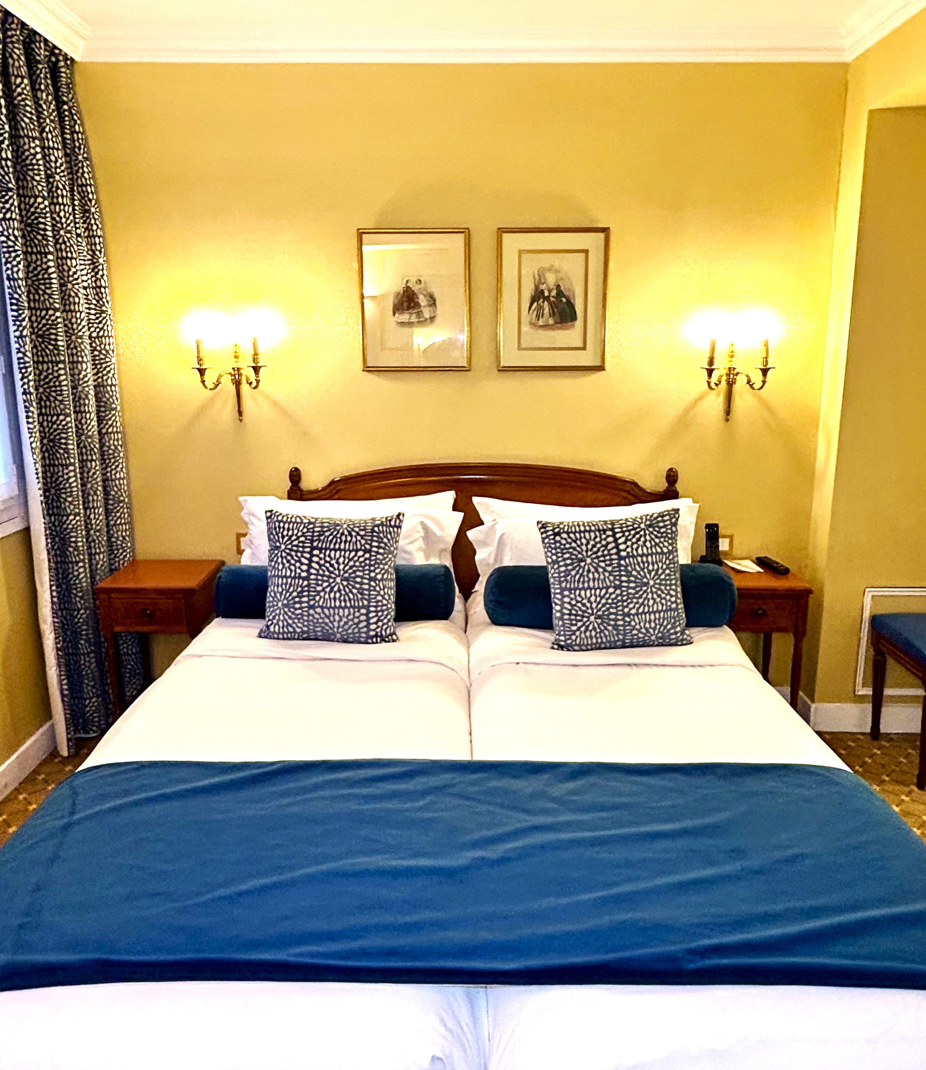 Een tweepersoonsbed met een blauwe spreideken, twee gestreepte kussens, een houten hoofdbord, twee wandlampen en twee ingelijste foto’s aan de muur.