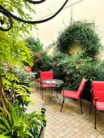 Un patio con sillas rojas y una mesa, rodeado de abundante vegetación y un suelo de baldosas.