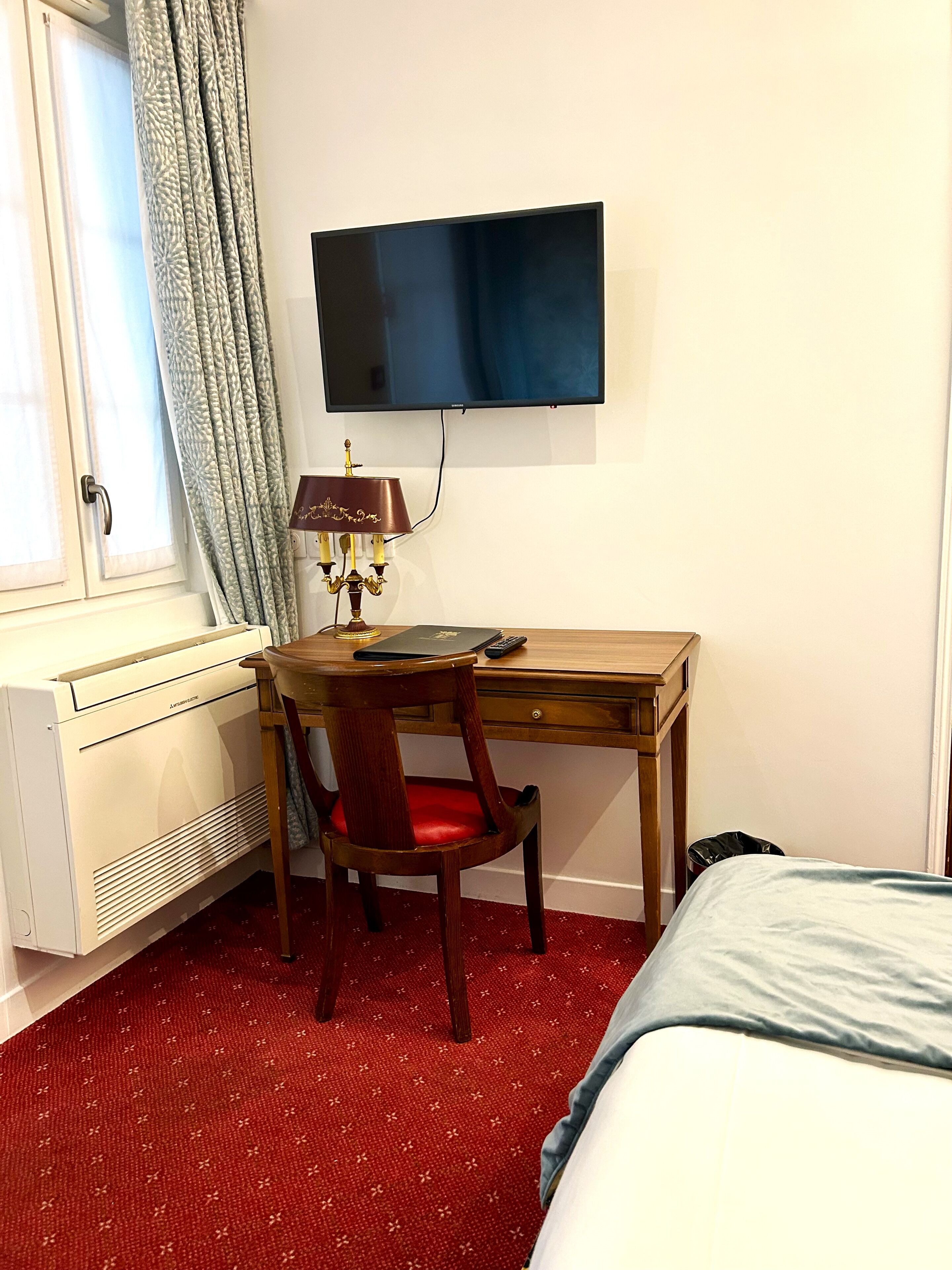 Une chambre d’hôtel avec un bureau en bois, une chaise rouge, un téléviseur fixé au mur, un radiateur et un lit recouvert d’une couverture bleue.