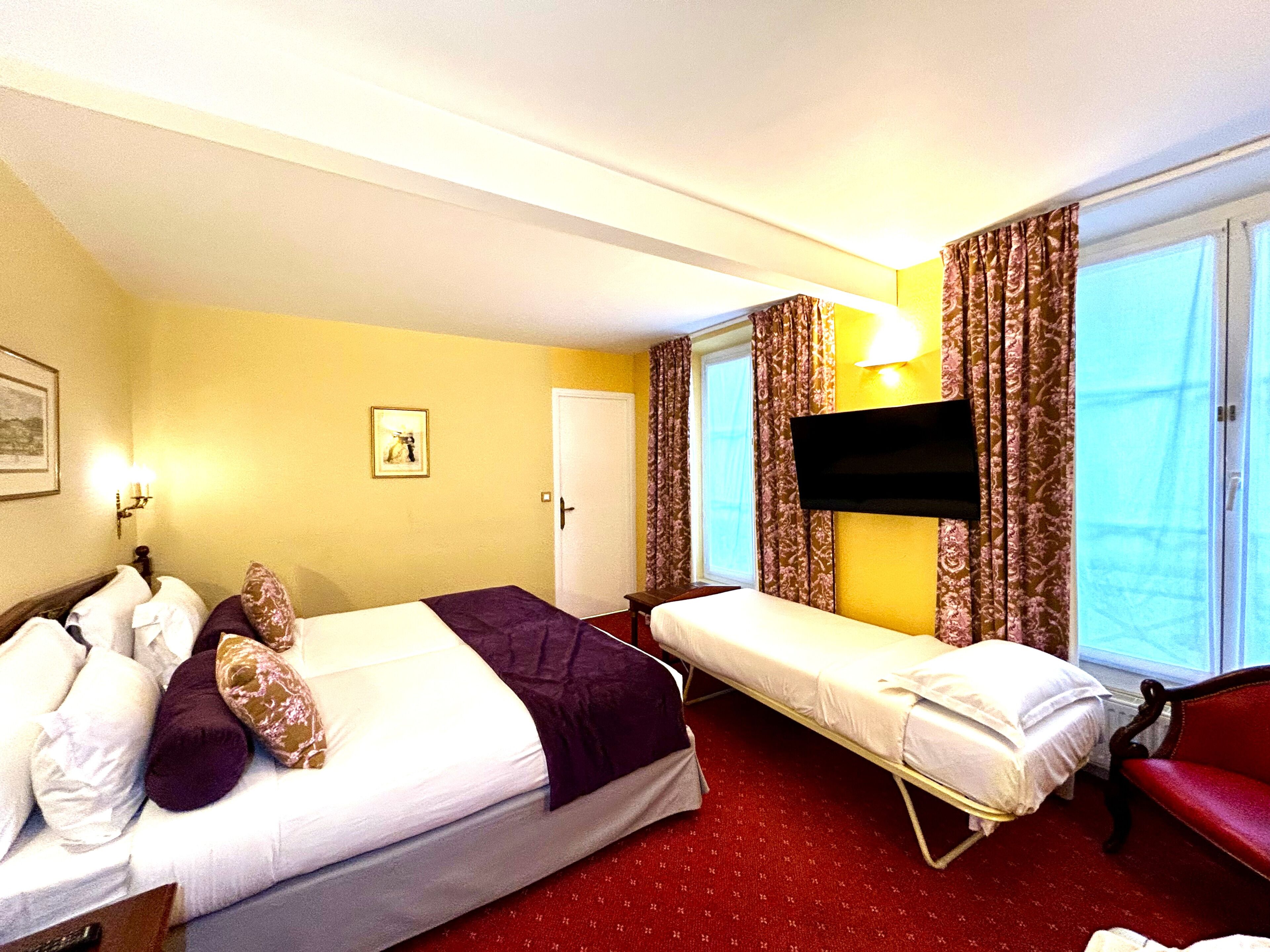 A hotel room with two beds, a TV, a red chair, a window with curtains, and a door.