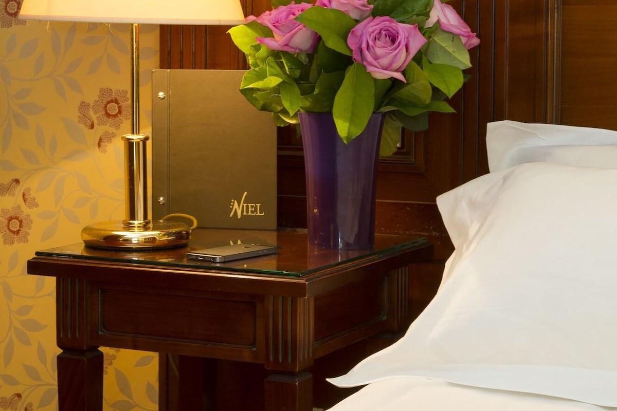 A hotel room with a bed, a nightstand with a lamp, a vase with flowers, and a sign with the hotel name.