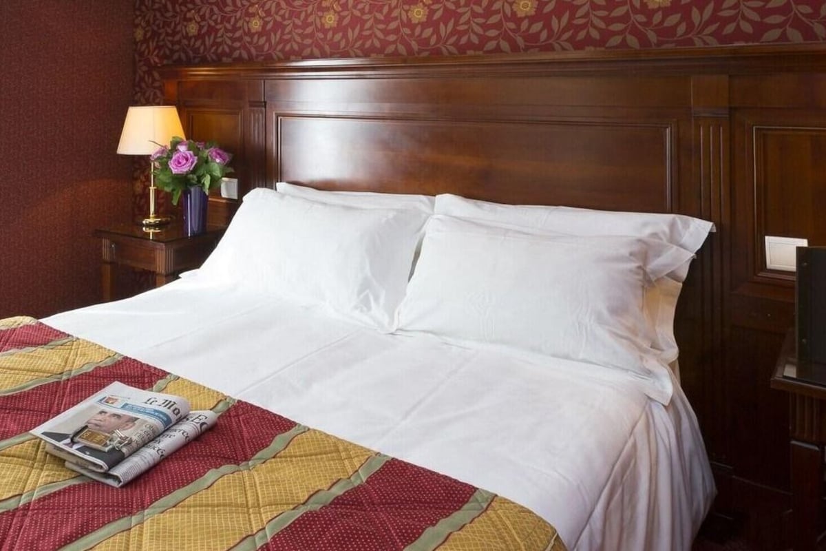 A hotel room with a neatly made bed, a floral-patterned wall, and a nightstand with a lamp and flowers.