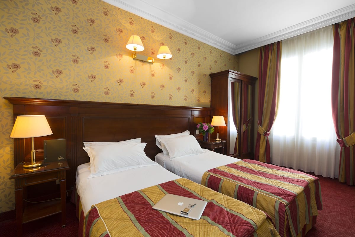Habitación de hotel con dos camas, cabecera de madera, mesita de noche con lámpara y ventana con cortinas.