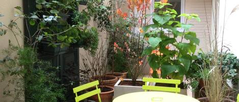 Un pequeño patio con una mesa redonda y dos sillas, rodeado de plantas en macetas y una puerta verde.