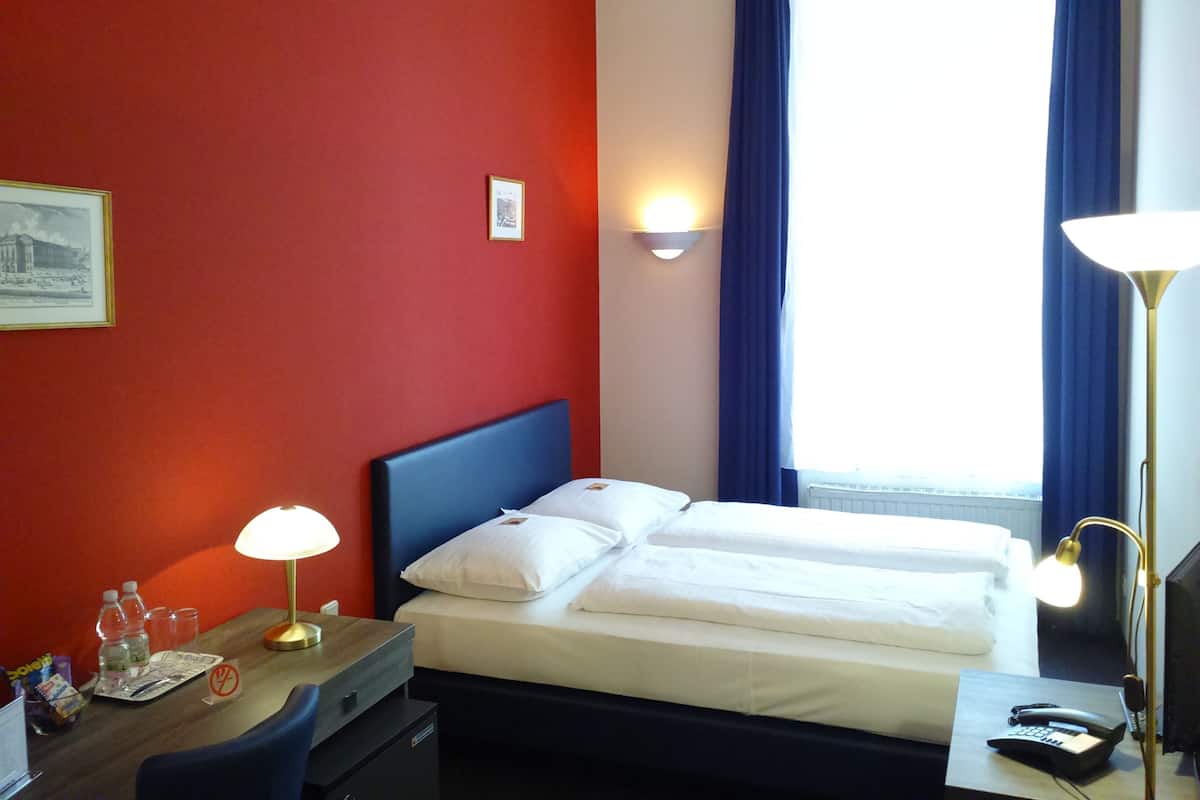Hotellihuone, jossa on sänky, yöpöytävalaisimet, kirjoituspöytä valaisimella, tuoli ja ikkuna, jossa on verhot.