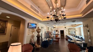A hotel lobby with a chandelier, dining area, and a TV displaying a beach scene. - Hotel Pension Excellence (Vienna)