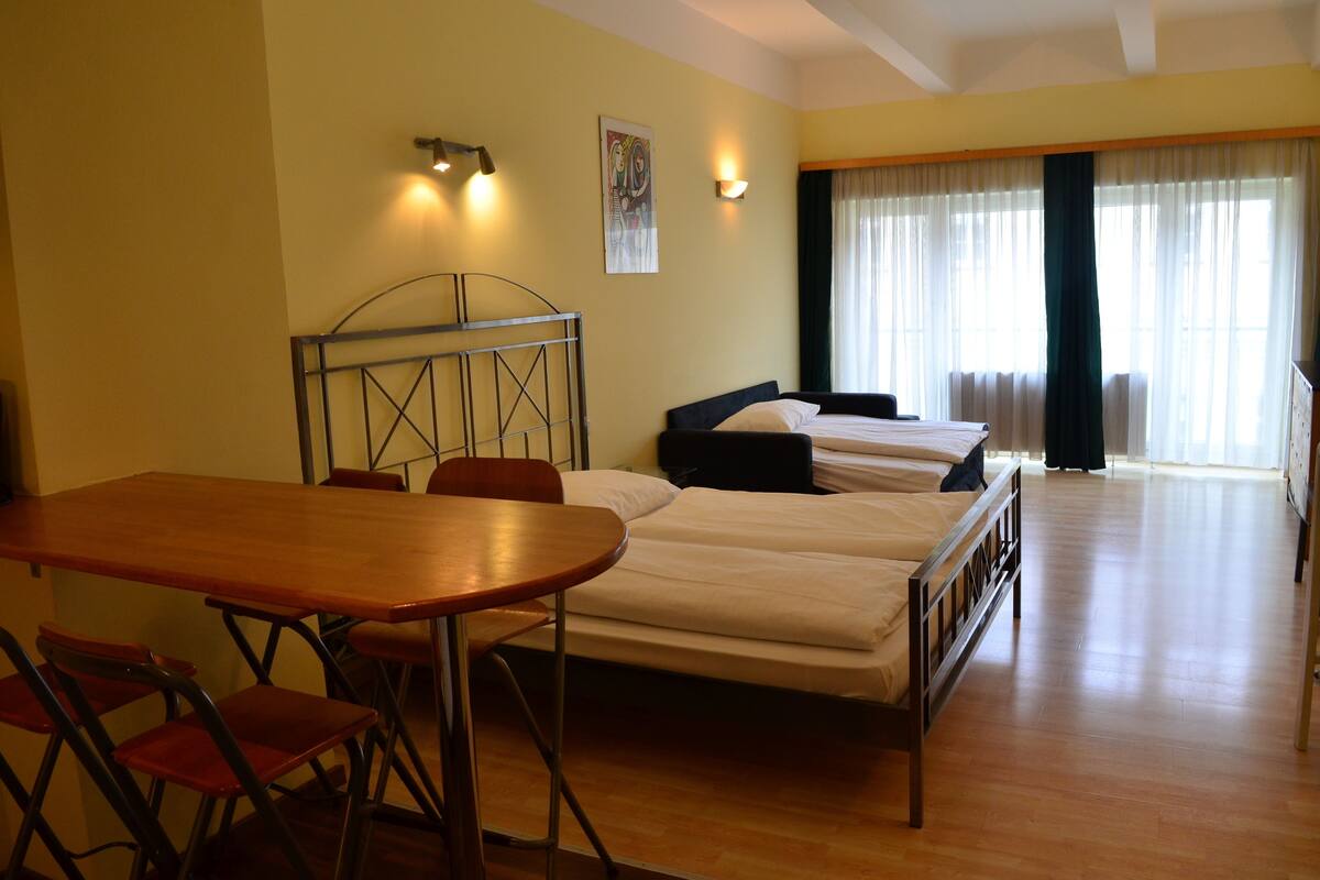 Hotellihuone, jossa on puinen pöytä ja tuolit, metallirungollinen sänky ja suuri ikkuna, jossa on verhot.