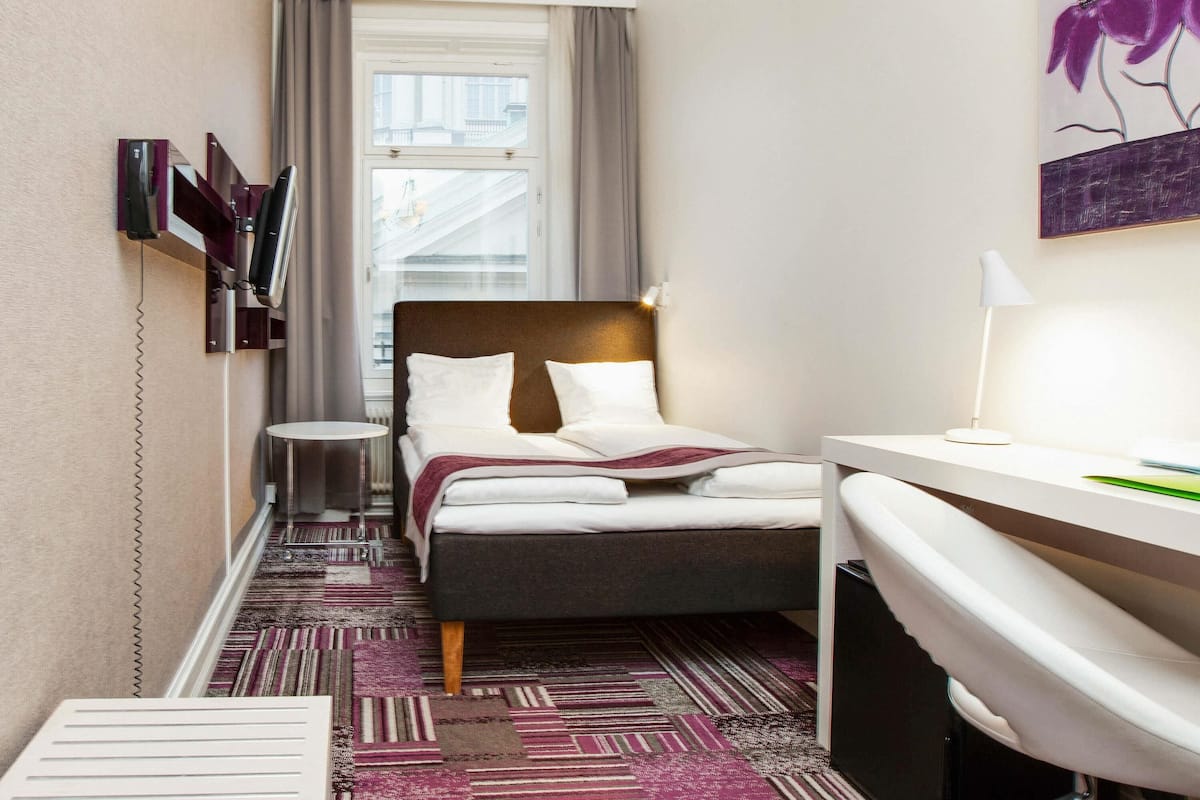 A hotel room with a bed, bedside table, TV, and a view of buildings outside the window.