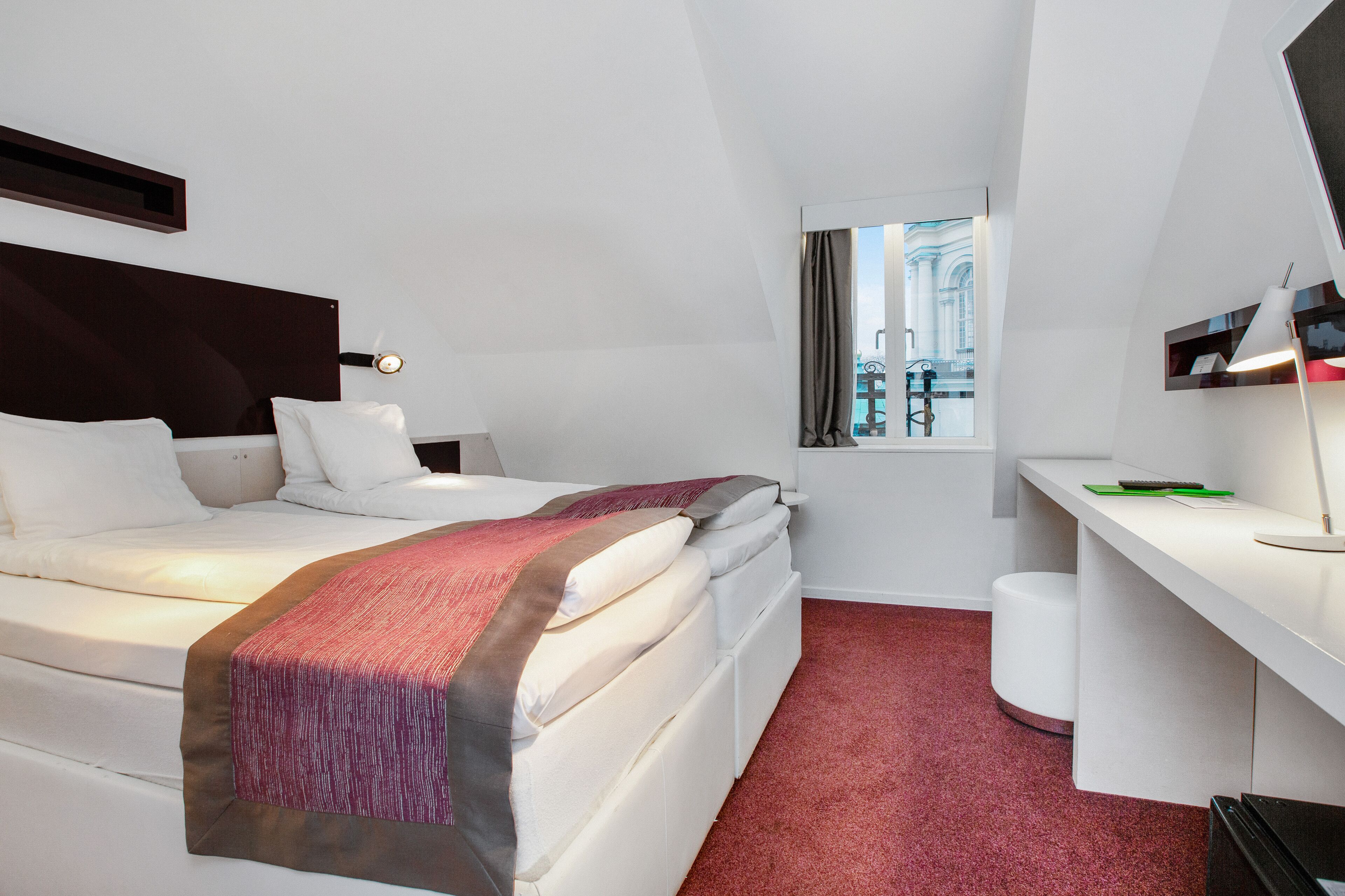 Une chambre d’hôtel comprenant un lit, un bureau, une chaise, une fenêtre avec des rideaux et une vue sur les bâtiments environnants.