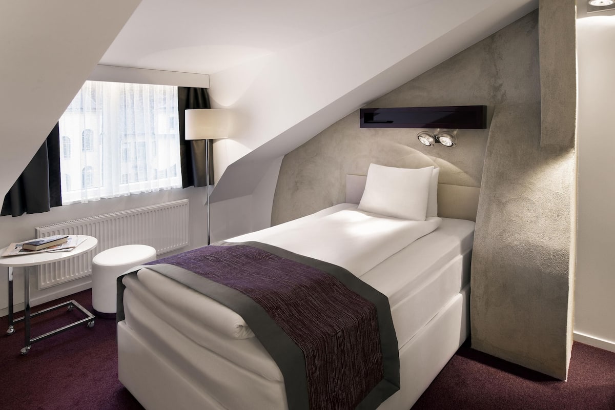 A modern hotel room with a single bed, a small table with a lamp, a radiator, and a view of the city through a window.