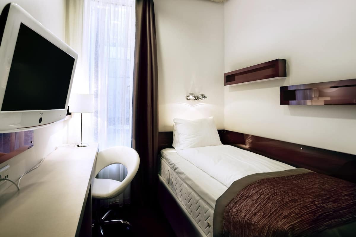A hotel room with a bed, a desk with a computer monitor, a chair, and a window with curtains.