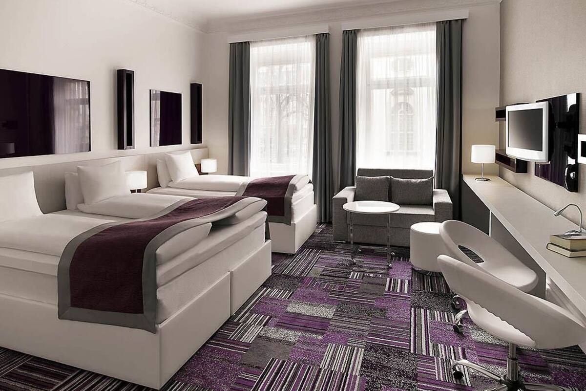 A hotel room with two beds, a desk, a chair, a TV, and large windows with curtains.