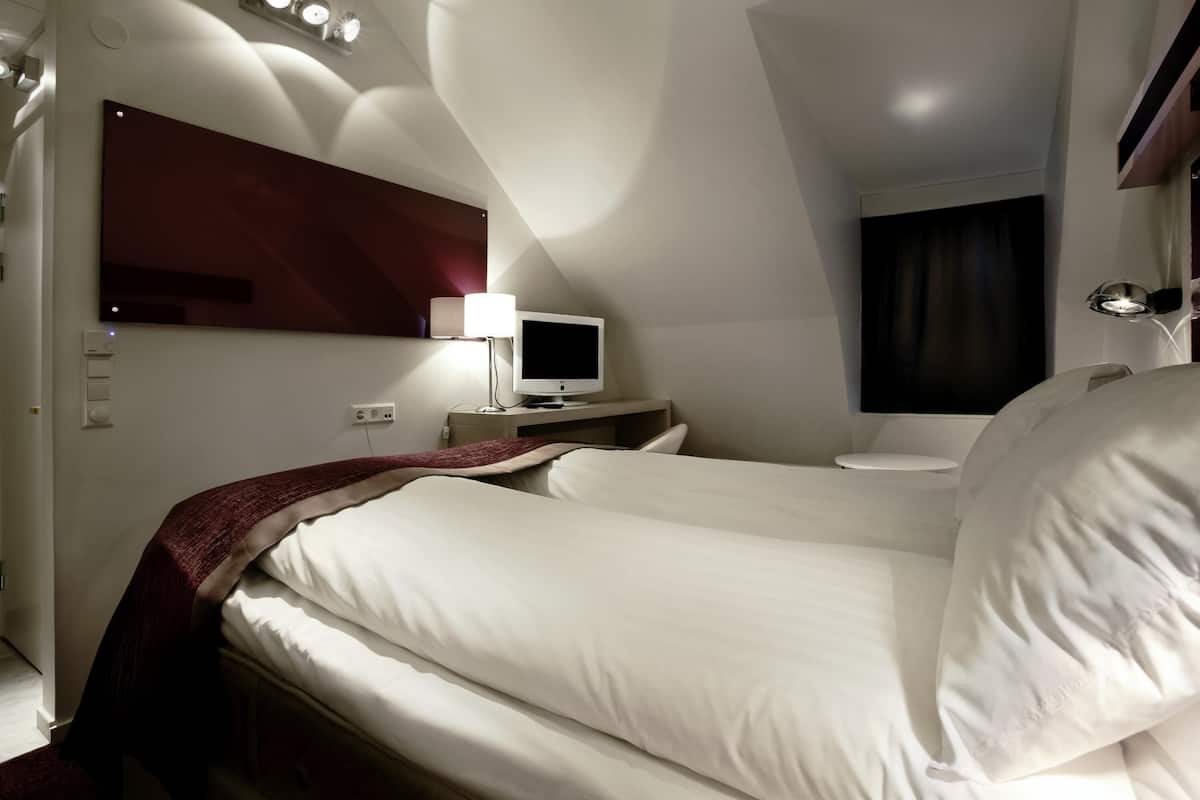 A hotel room with a bed, a TV, a bedside table, a lamp, a mirror, and a wall-mounted clock.