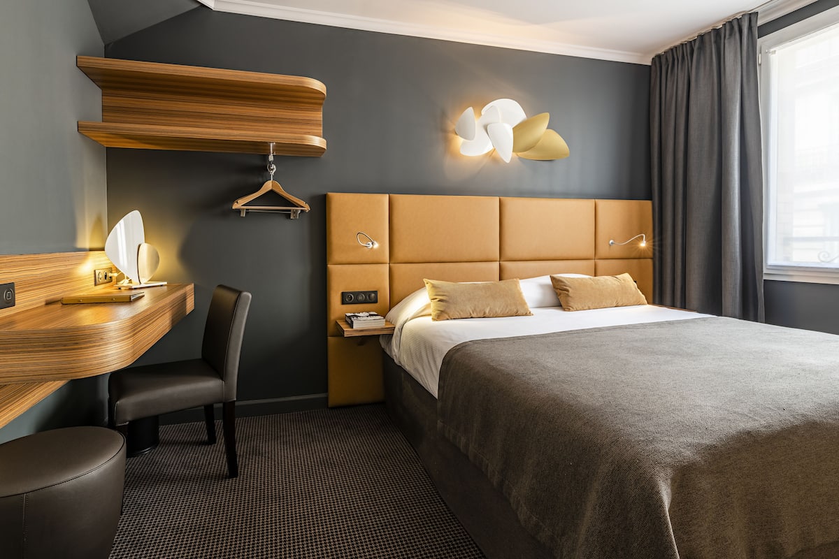 A modern hotel room with a large bed, a desk with a chair, a hanging coat rack, and a wall-mounted light fixture.