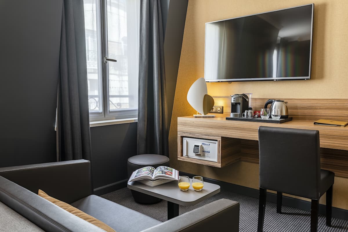 A modern hotel room with a sofa, a small table with a book and glasses, a desk with a coffee maker, a TV, and a window with curtains.