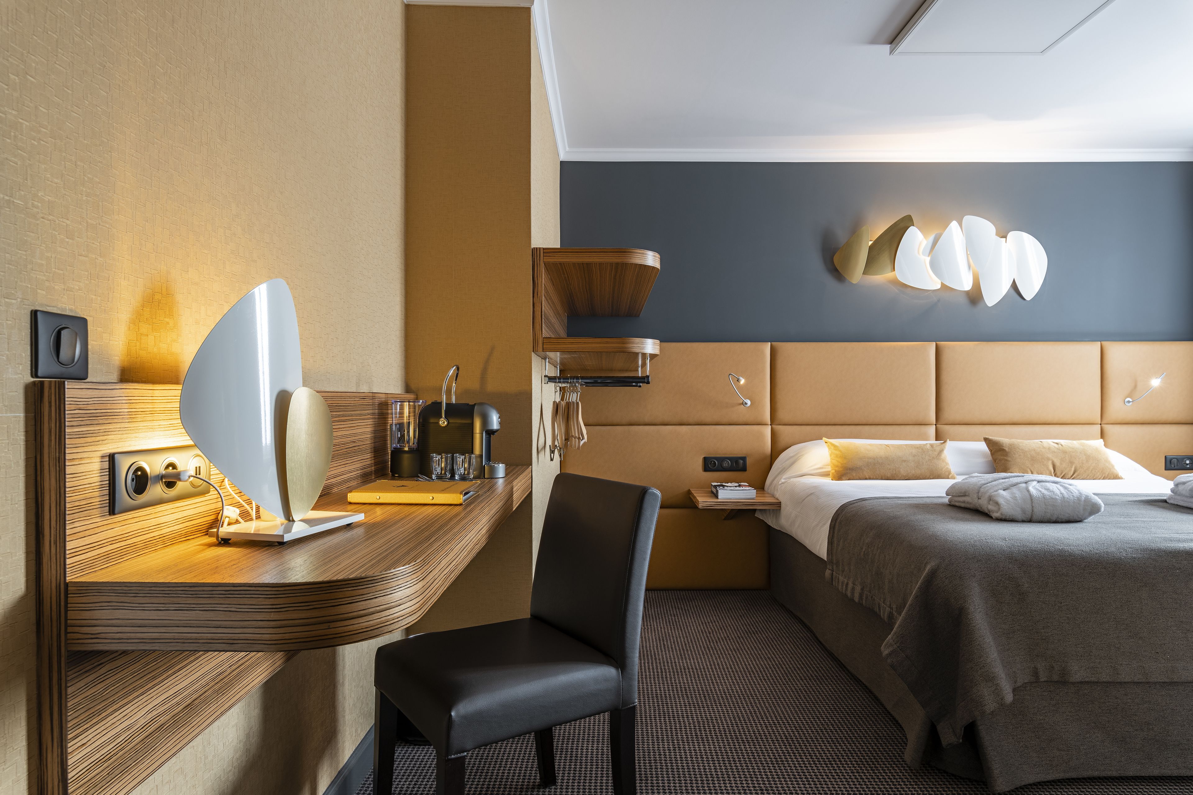 A hotel room with a modern design, featuring a large bed with a headboard, a desk with a chair, a wall-mounted lamp, and a bedside table with a lamp.