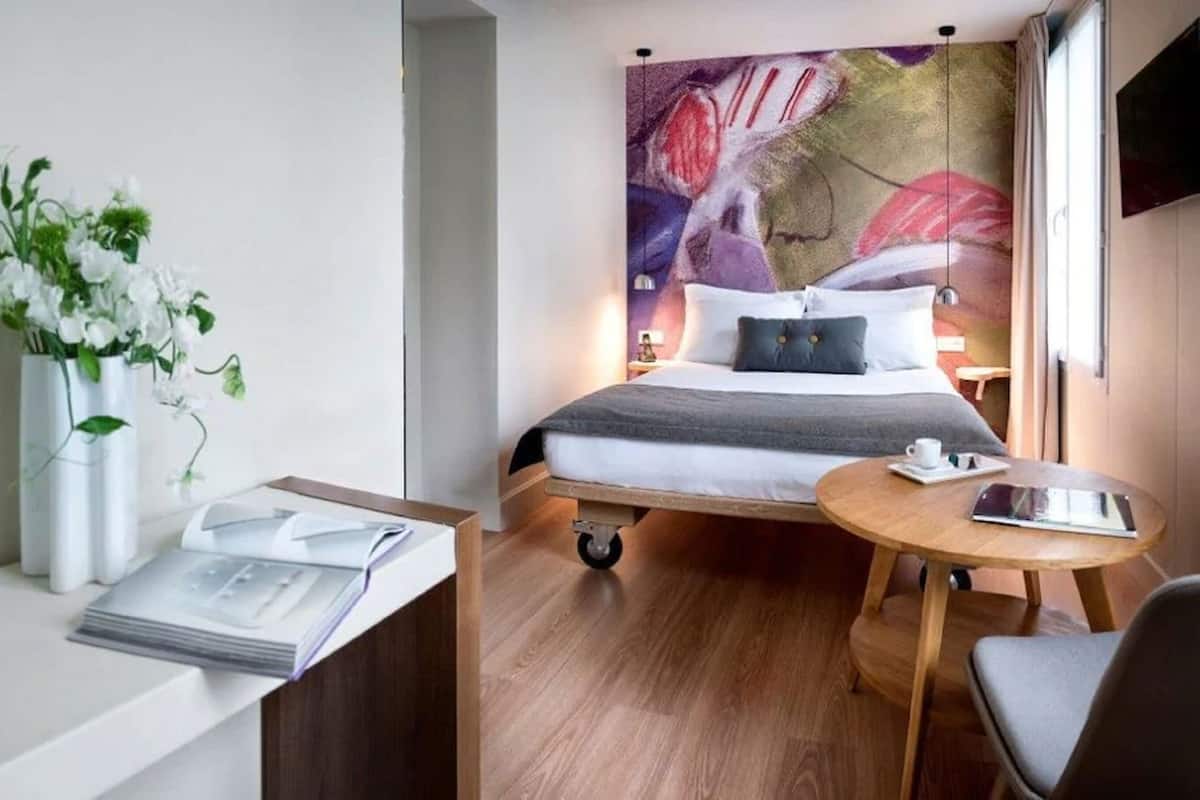 A hotel room with a bed, a desk with a chair, a small table, and a wall with a floral painting.