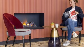 A person sitting on a checkered chair with a red velvet backrest, reading a book. There's a small table with a plant and a cup, and a fireplace with a fire in the background. - Hotel de la Paix (Paris)