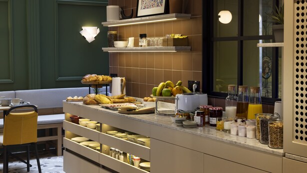 A breakfast bar with various food items and beverages, a seating area with a bench, and a window with a view. - Hotel de la Paix (Paris)