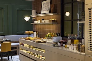 A breakfast bar with various food items and beverages, a seating area with a bench, and a window with a view. - Hotel de la Paix (Paris)