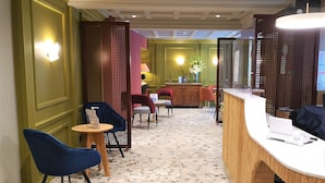 A reception area with a wooden reception desk, blue chairs, a small round table, and a decorative wall with perforated panels. - Hotel de la Paix (Paris)