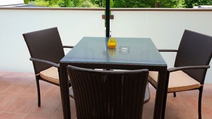 A patio with a glass table and wicker chairs, a glass of orange juice, and a clear view of a green garden and trees. - Hotel Via Roma (Salzburg)
