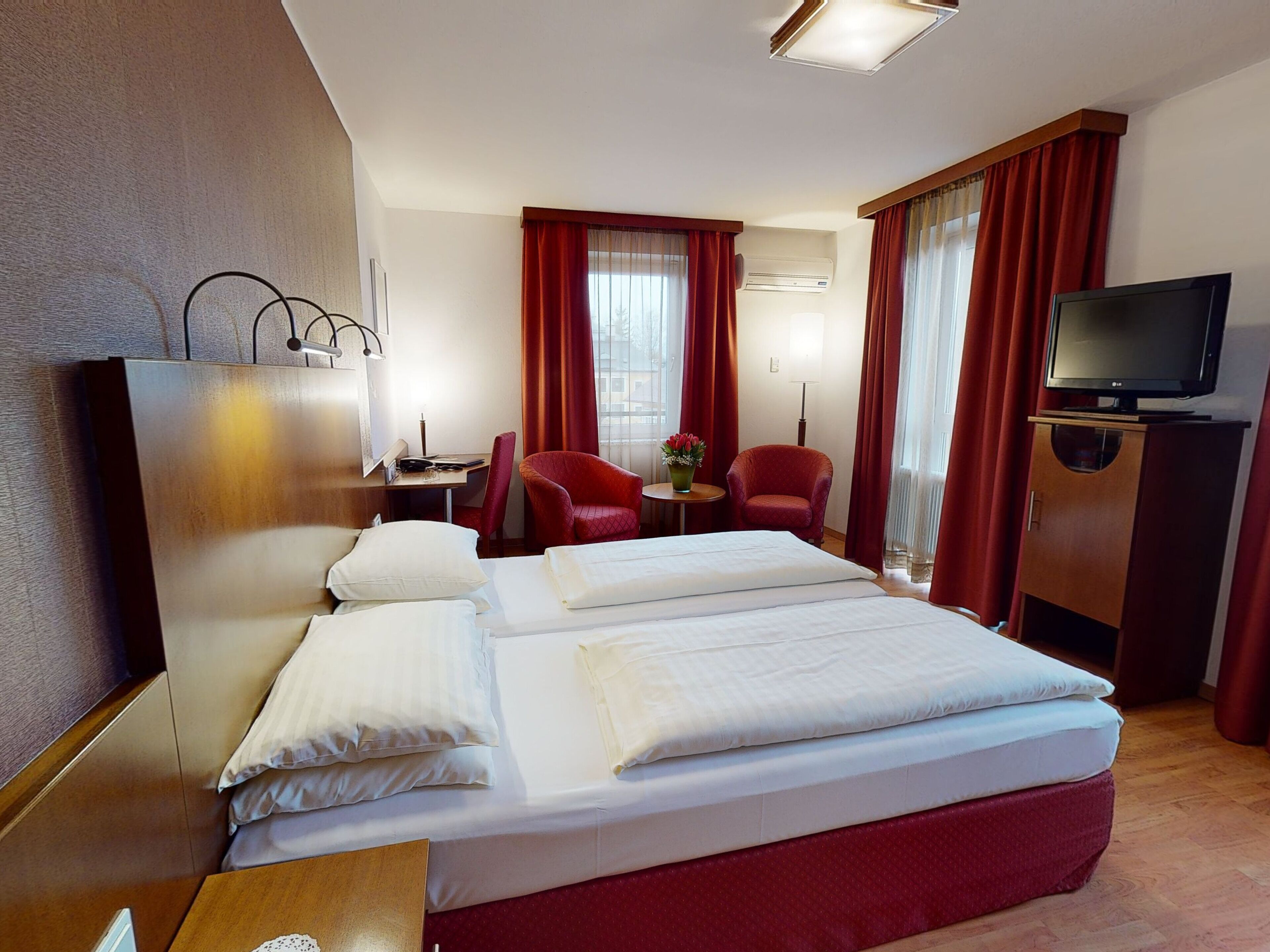 A hotel room with a bed, two red chairs, a TV, and a desk with a lamp.