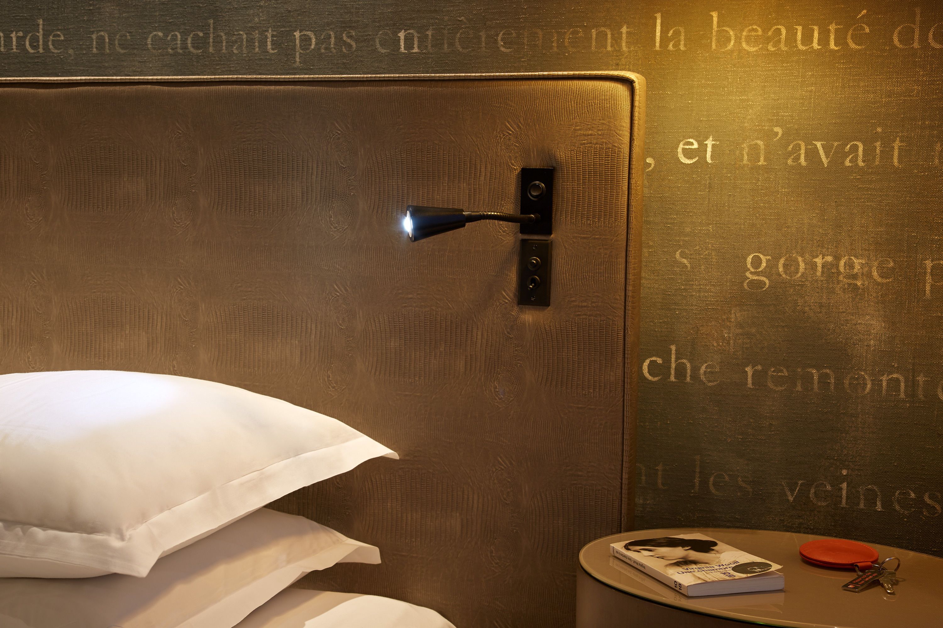 A hotel room with a bed, pillows, a bedside table with a book, a remote control, and a wall with French text.